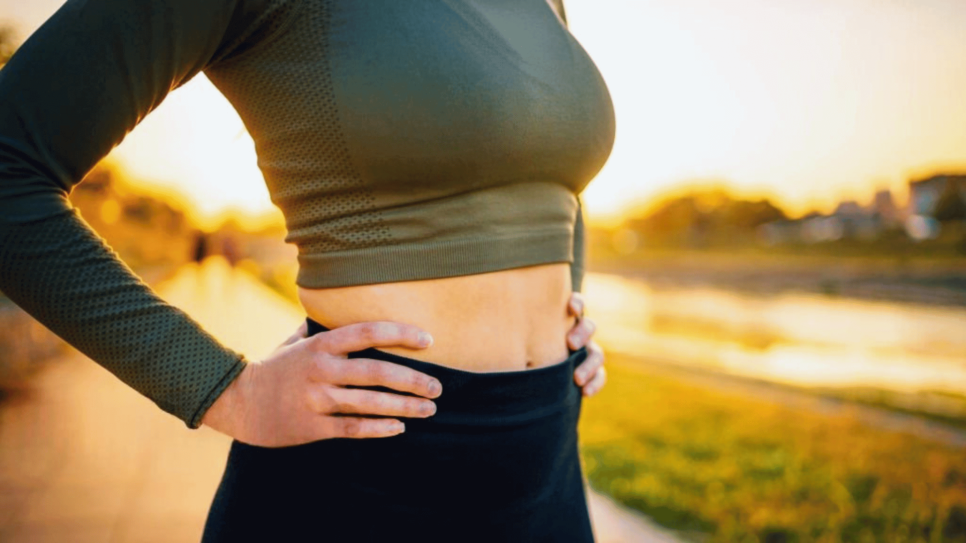 Femme en tenue de sport, mains sur les hanches, montrant son ventre plat au coucher du soleil, symbole de bien-être et de remise en forme.