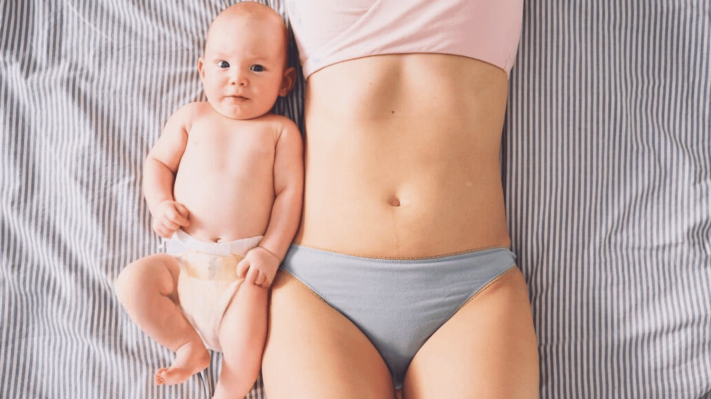 Jeune mère allongée en sous-vêtements à côté de son bébé en couche, montrant son ventre plat après l’accouchement.