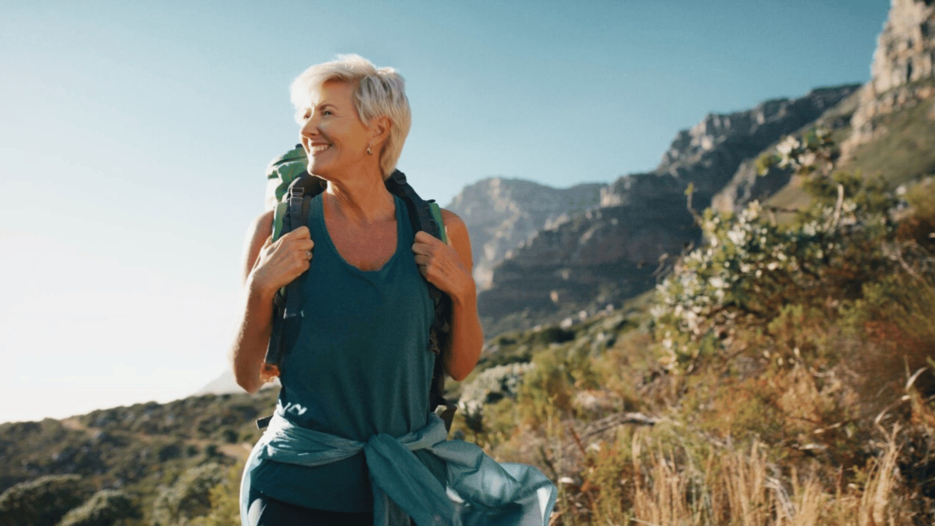 Femme senior souriante en randonnée, portant un sac à dos et regardant vers l’horizon, avec des montagnes en arrière-plan sous un ciel bleu clair.