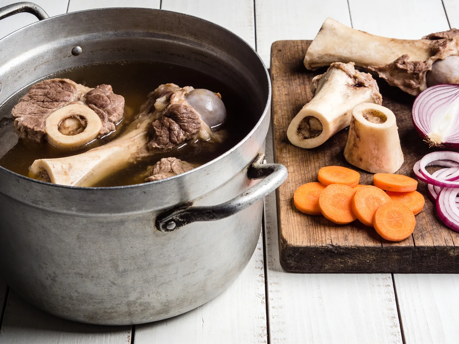 Marmite en aluminium remplie de bouillon avec des os et de la viande de bœuf, à côté d'une planche en bois avec os à moelle, carottes et oignon rouge