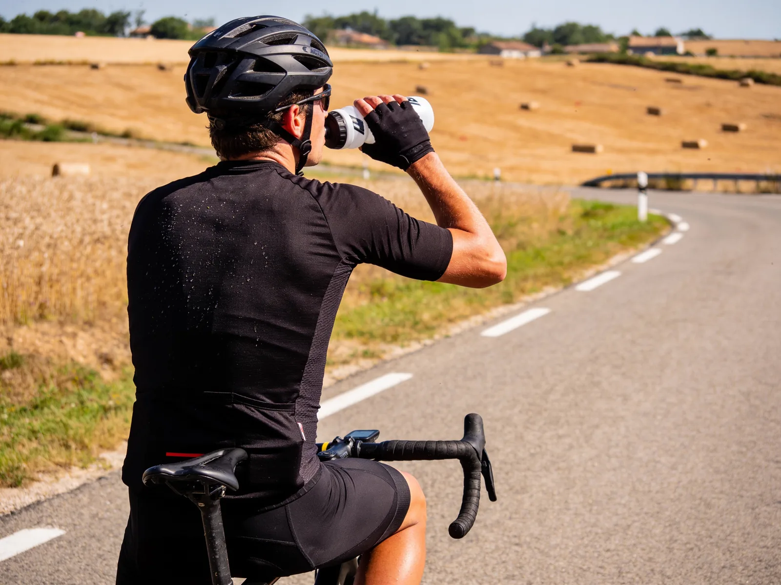 Cycliste vu de dos sur une route de campagne qui boit à son bidon, maillot noir marqué de sueur, avec des champs secs et des bottes de paille derrière lui