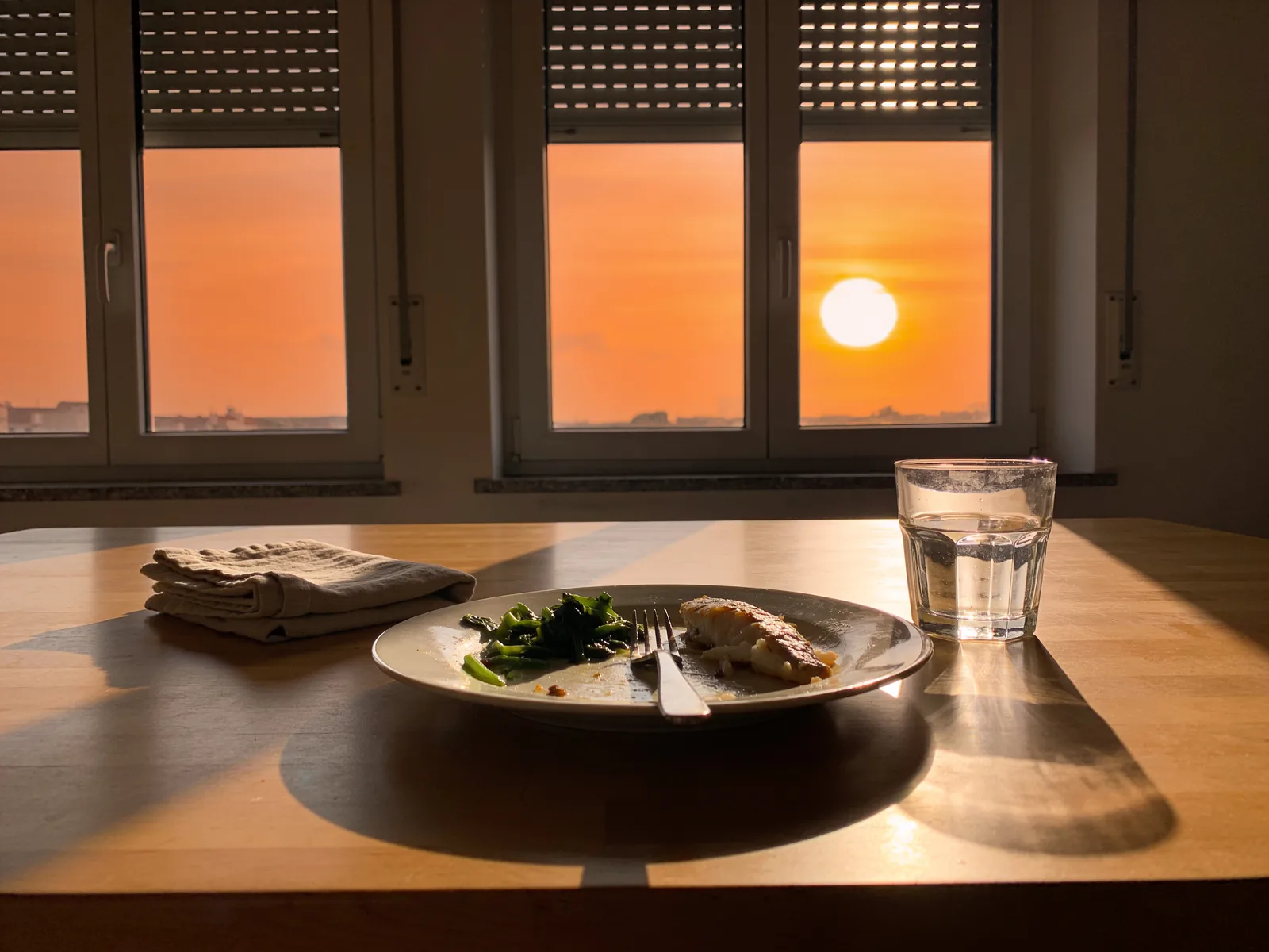Restes de poisson et de légumes verts dans une assiette sur une table en bois, avec serviette en lin et verre d'eau, au coucher du soleil